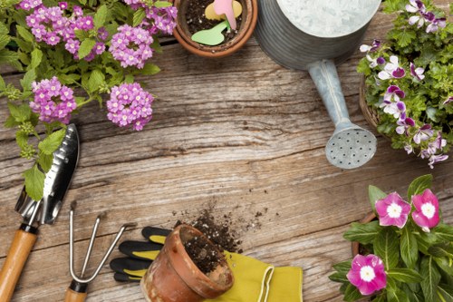 A top-down view of a wooden garden table displaying gardening tools and materials, including a small garden fork, a hand rake, a pair of gardening gloves, and a terracotta pot with soil. Surrounding the tools, there are three flower pots filled with vibrant pink and purple flowering plants, creating a colourful outdoor scene that suggests ongoing gardening activity in a residential garden or backyard, possibly in Finchley, London. The weather appears to be mild and suitable for outdoor planting or maintenance, aligning with gardening services offered by Landscapers Finchley. The natural textures of the weathered wood and the bright, healthy foliage of the flowers highlight a well-maintained garden space.