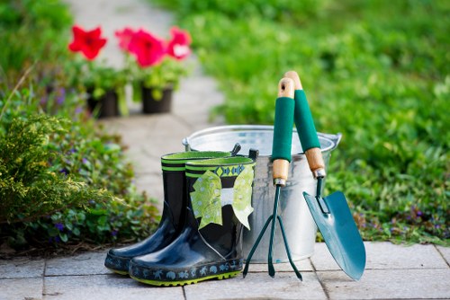 A pair of black rubber gardening boots with green patterned socks inside are placed on a paved outdoor area in a garden. Next to the boots, there is a white plastic bucket, a small hand fork, and a trowel with green handles resting against the bucket. In the background, vibrant pink and red flowering plants are visible, situated along a narrow pathway that leads through a lush, green garden space. The garden features neatly maintained grass, flowering shrubs, and a variety of foliage, suggesting a well-cared-for outdoor area designed for gardening activities. The scene is illuminated by natural daylight with clear weather, highlighting the tools and plant life, and exemplifying typical garden maintenance elements that a landscaping company like Landscapers Finchley might focus on for enhancing outdoor spaces in Finchley or nearby areas.