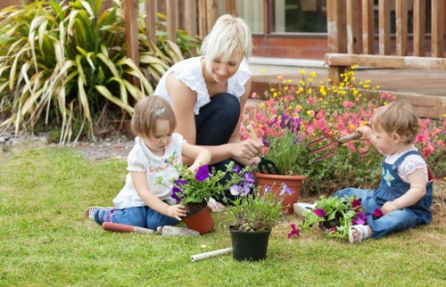A woman with short blonde hair, dressed in a white top with short sleeves, is seated on a well-maintained, lush green lawn in a garden, surrounded by two young girls with light brown hair wearing casual clothes, all engaged in planting and tending to various potted flowers, including purple and pink blooms. The garden features a backdrop of ornamental grasses with long, pointed leaves, a wooden deck or patio area with steps leading down to the lawn, and a variety of colorful flowering plants in flower beds close to the house, which has large windows visible behind the scene. The scene is lit with natural daylight, suggestive of a bright, clear day, with an overall impression of neatness, outdoor leisure, and garden care, aligning with local lawn and garden maintenance services in Finchley.
