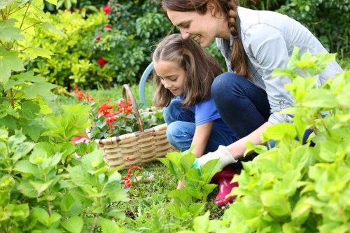A woman and a young girl are kneeling in a lush garden with dense, green foliage and vibrant flowering plants, including red and pink blooms, surrounded by leafy shrubs and trees. The woman, with long brown hair tied back, is wearing a light grey jacket and blue jeans, while the girl, with brown hair and a blue shirt, is holding a wicker basket and appears to be picking flowers or plants. The garden has a well-maintained lawn area with dense grass, bordered by flower beds, and a paved patio or pathway visible in the background, suggesting an outdoor residential garden typical of Finchley area. The scene is set in natural daylight, indicating fair weather with no signs of rain or harsh sunlight, highlighting the healthy, verdant condition of the plants and the pleasant outdoor environment that landscaping and gardening services by Landscapers Finchley aim to enhance and maintain.