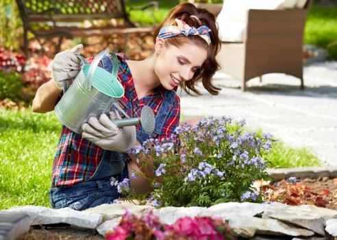 A young woman with light skin, wearing a multicoloured checkered shirt, grey gardening gloves, and a floral headband, is kneeling in a neatly maintained garden in Finchley. She is smiling as she waters a cluster of small, vibrant purple and light blue flowering plants with a metal watering can. The garden features a well-defined stone-edged flower bed with colorful plants, a lush green lawn in the foreground, and a background of wooden garden furniture and a paved patio area. Behind her, there are dense, green hedges and trees, with sunlight illuminating the scene and casting subtle shadows, suggesting a pleasant, mild weather day. The setting reflects a typical suburban outdoor space in North London, suitable for gardening and outdoor maintenance services provided by Landscapers Finchley, with attention to planting details, garden layout, and overall landscape care.