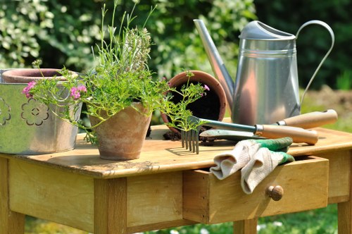 A wooden garden table outdoors in a lush green garden setting, displaying various gardening tools and accessories such as a silver watering can, a pair of garden gloves, and several flower pots filled with vibrant green plants and delicate pink flowers. The table surface shows signs of gardening activity, with some soil visible in the pots and tools arranged for use. In the background, dense foliage and trees suggest a well-maintained backyard garden, providing a natural and inviting environment for gardening tasks. The lighting indicates a bright day, highlighting the textures of the wood, plants, and gardening equipment. This scene exemplifies typical gardening activities that a professional landscapers Finchley company such as landscapersfinchley.co.uk offers, including planting, potting, and garden maintenance in a residential outdoor space near Finchley.