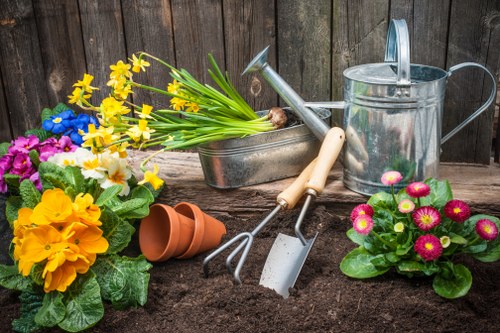 A neatly maintained backyard garden with a durable wooden fence in the background, featuring a landscaped flower bed with dark, rich soil in the foreground. The bed contains vibrant flowering plants, including bright yellow daffodils, pink primroses, and purple pansies, alongside green foliage. Nearby, gardening tools such as a small trowel, a hand rake, and a watering can are arranged on the soil, indicating recent planting or maintenance activity. To the side, two upside-down terracotta pots sit on the ground, suggesting future plantings or repurposing. The outdoor space is well-organized, with natural daylight illuminating the lush, healthy plants and freshly turned soil, reflecting professional gardening and landscaping care in Finchley, in the London area. This scene supports gardening services that include flower planting, soil preparation, and outdoor garden maintenance, tailored to the local area’s climate and garden styles.