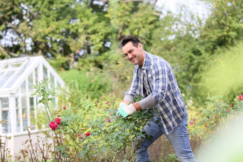 A man in casual clothing and gardening gloves is tending a flower bed in a well-maintained garden, surrounded by various plants, shrubs, and blooming flowers. His attention is focused on the vibrant pink and red flowers, with a greenhouse structure visible in the background on the left side, indicating a cultivated outdoor space in Finchley. The garden features a lush, green lawn area contrasting with the darker soil of the flower beds, and is bordered by hedges and trees that provide natural privacy and shade. The weather appears bright and mild, with natural sunlight illuminating the scene, emphasizing the vivid colours and textures of the foliage and blossoms. This setting aligns with services offered by Landscapers Finchley in gardening and landscaping, highlighting the importance of garden maintenance, planting, and outdoor aesthetic enhancement in the Finchley area. The environment suggests a residential back garden, characterized by a harmonious balance of cultivated vegetation and open grassed areas, suitable for outdoor leisure and gardening activities.