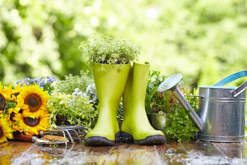 The image features a garden scene with a wooden outdoor table holding gardening essentials and decorative plants in a backyard setting. Prominently, a pair of bright green rubber rain boots are used as plant holders, filled with small white flowers and placed centrally on the table. To the left of the boots, there is a bouquet of sunflowers mixed with other purple and yellow flowers, adding vibrant colour to the scene. On the right side, a silver metal watering can with a blue handle is positioned, with a small spout and a detachable spray head. The background showcases lush green foliage and trees, with dappled sunlight filtering through the leaves, creating a fresh and natural outdoor environment. The surface of the table appears slightly wet, hinting at recent watering or rain. The overall composition emphasizes a well-maintained garden space suitable for gardening tasks and outdoor leisure, reflecting the services offered by Landscapers Finchley in garden and outdoor area improvements around Finchley, N12.